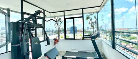A modern gym with a treadmill, weight machine, and a bench. It has large windows offering a city view.