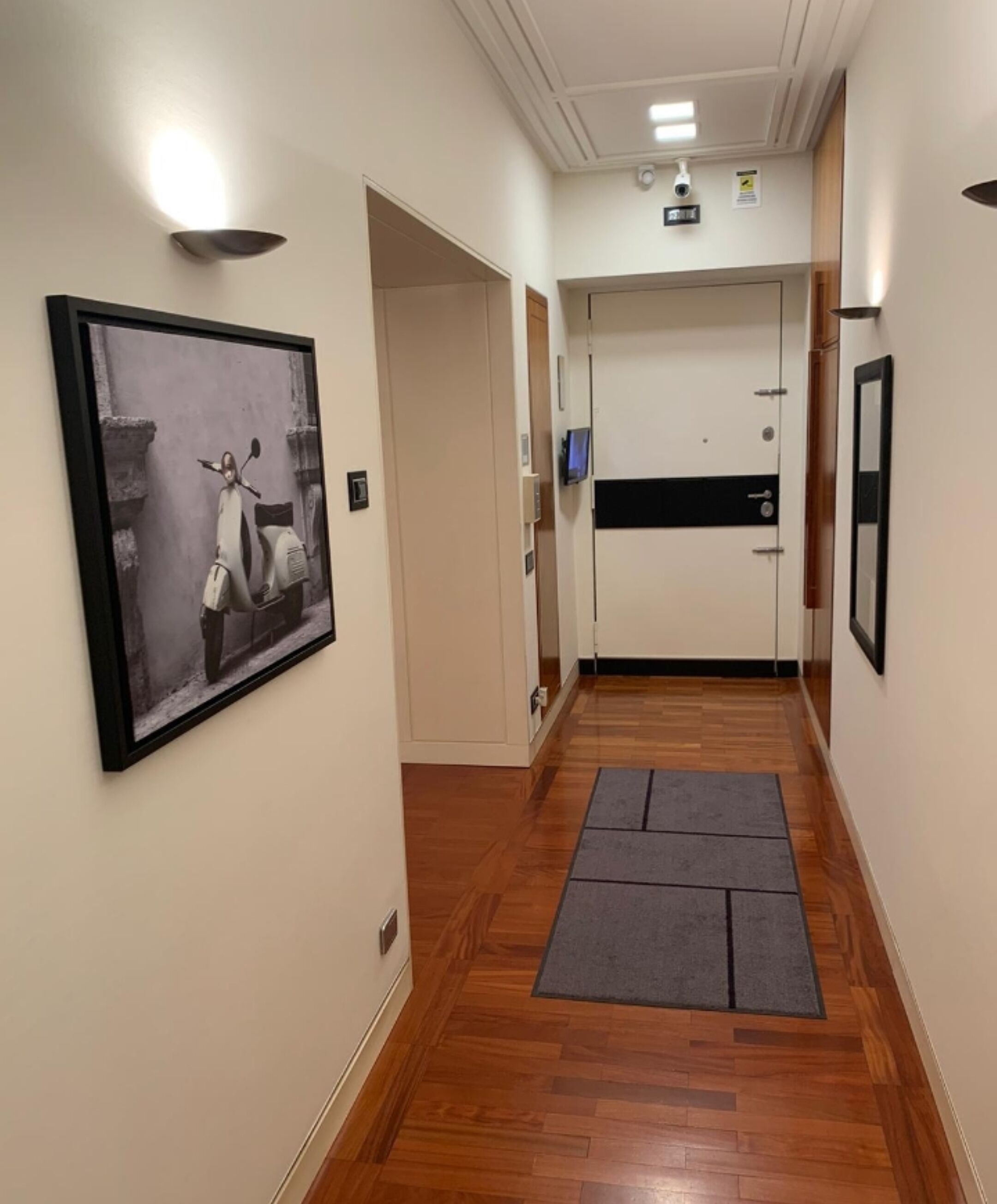 Un corridoio con pavimento in legno, una foto incorniciata al muro, una porta con una striscia nera e una telecamera di sicurezza a soffitto.