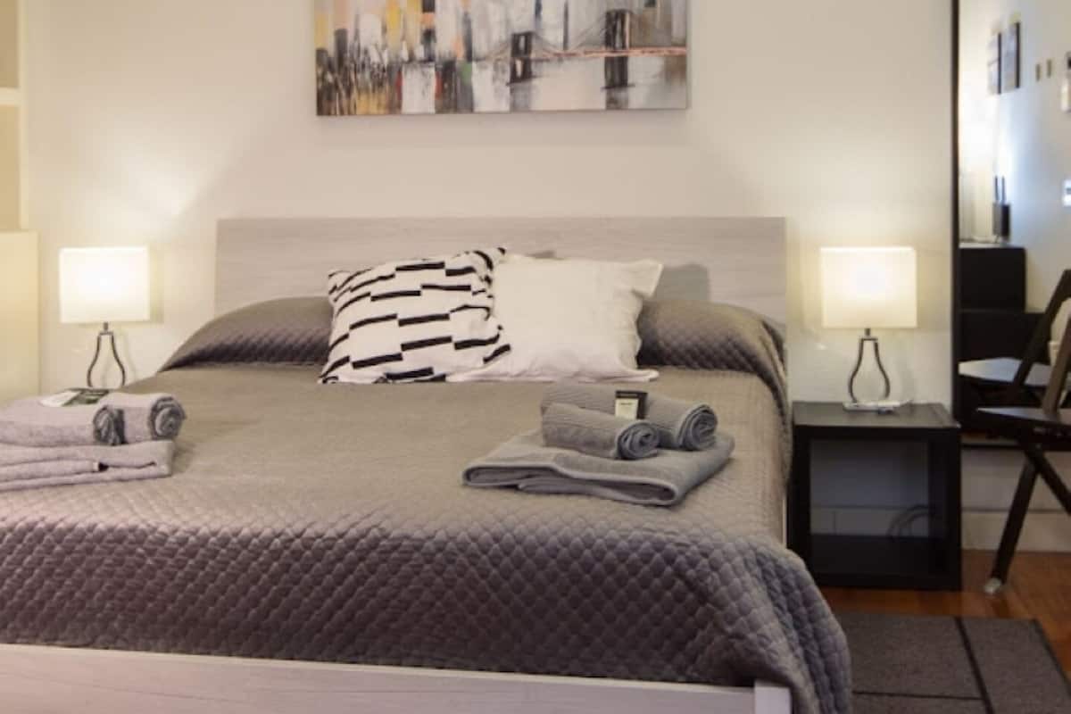 Un lit bien fait, avec une couette grise matelassée, deux oreillers et des serviettes enroulées. Un tableau est accroché au mur au-dessus du lit, et une table de chevet avec une lampe se trouve à droite.