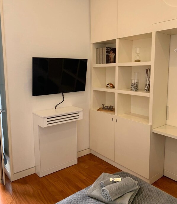 A bedroom with a TV mounted on a white stand, a wardrobe with open shelves, and a bed with a quilted cover. - b&b mimmù (Naples)