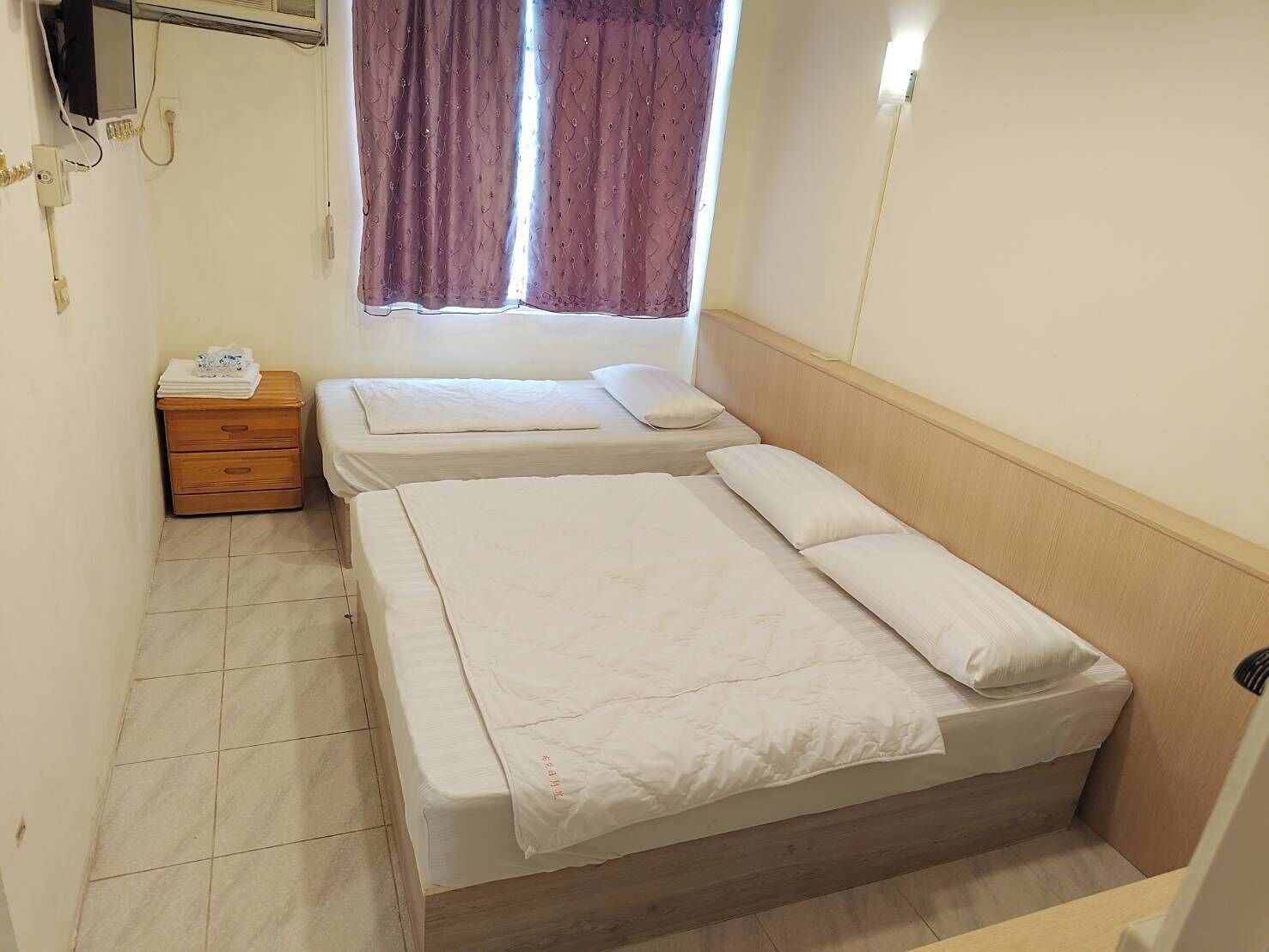 A small, clean room with two single beds, a wooden nightstand, a mirror, and a window with curtains.