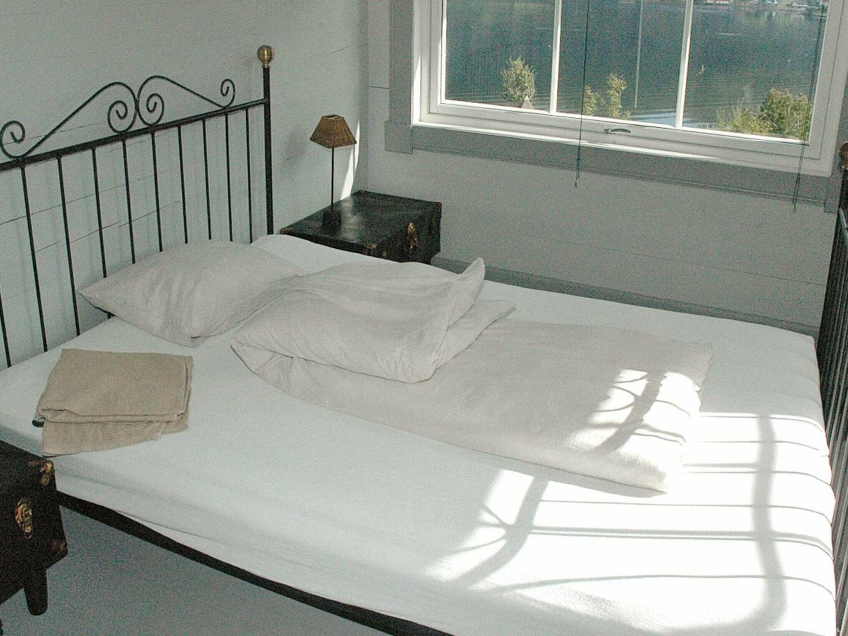 A single bed with white linens, a bedside table with a lamp, and a window with a view of trees.