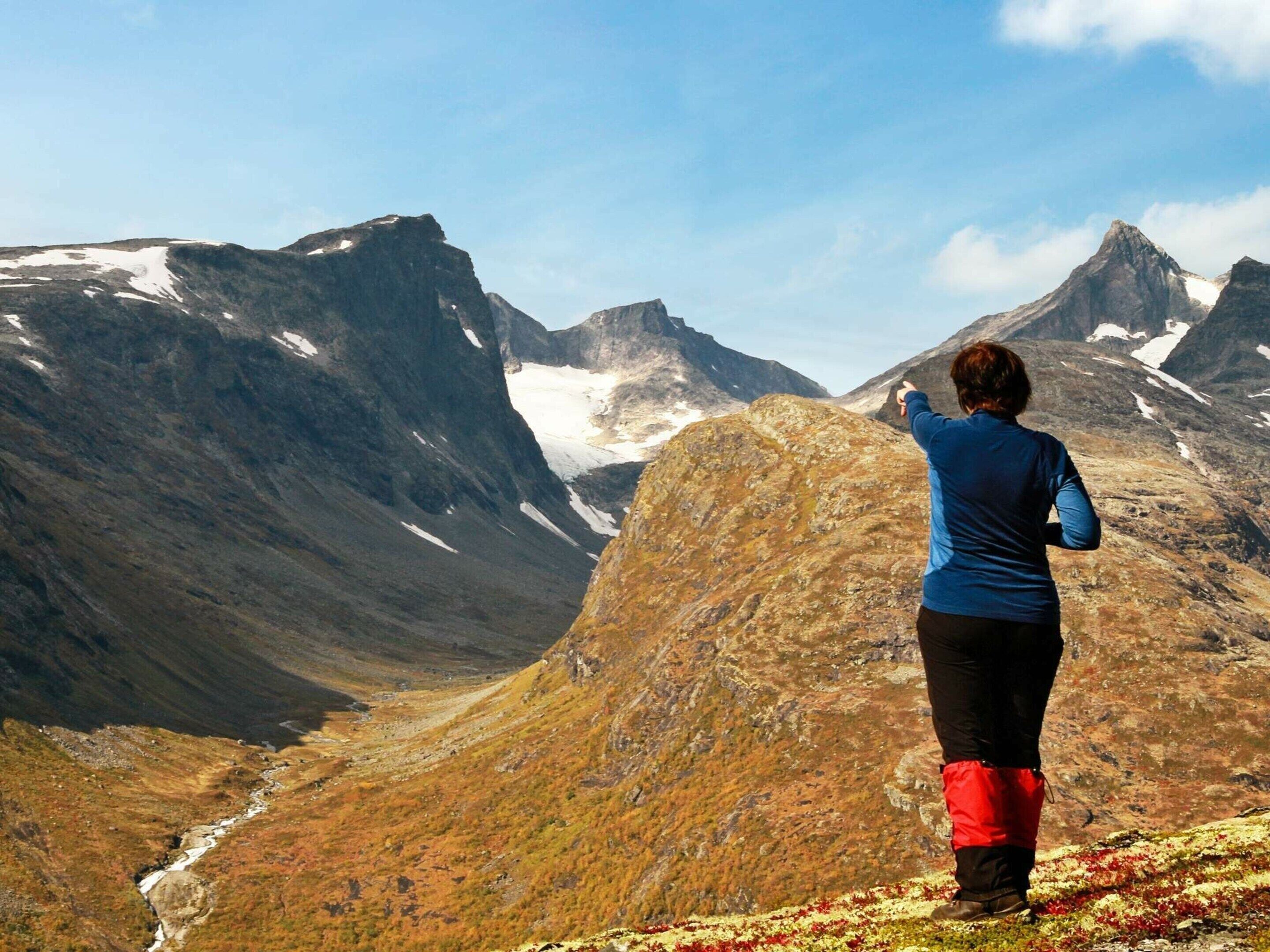 Una persona en la cresta de una montaña, con vista a un valle con parches de nieve y un río.