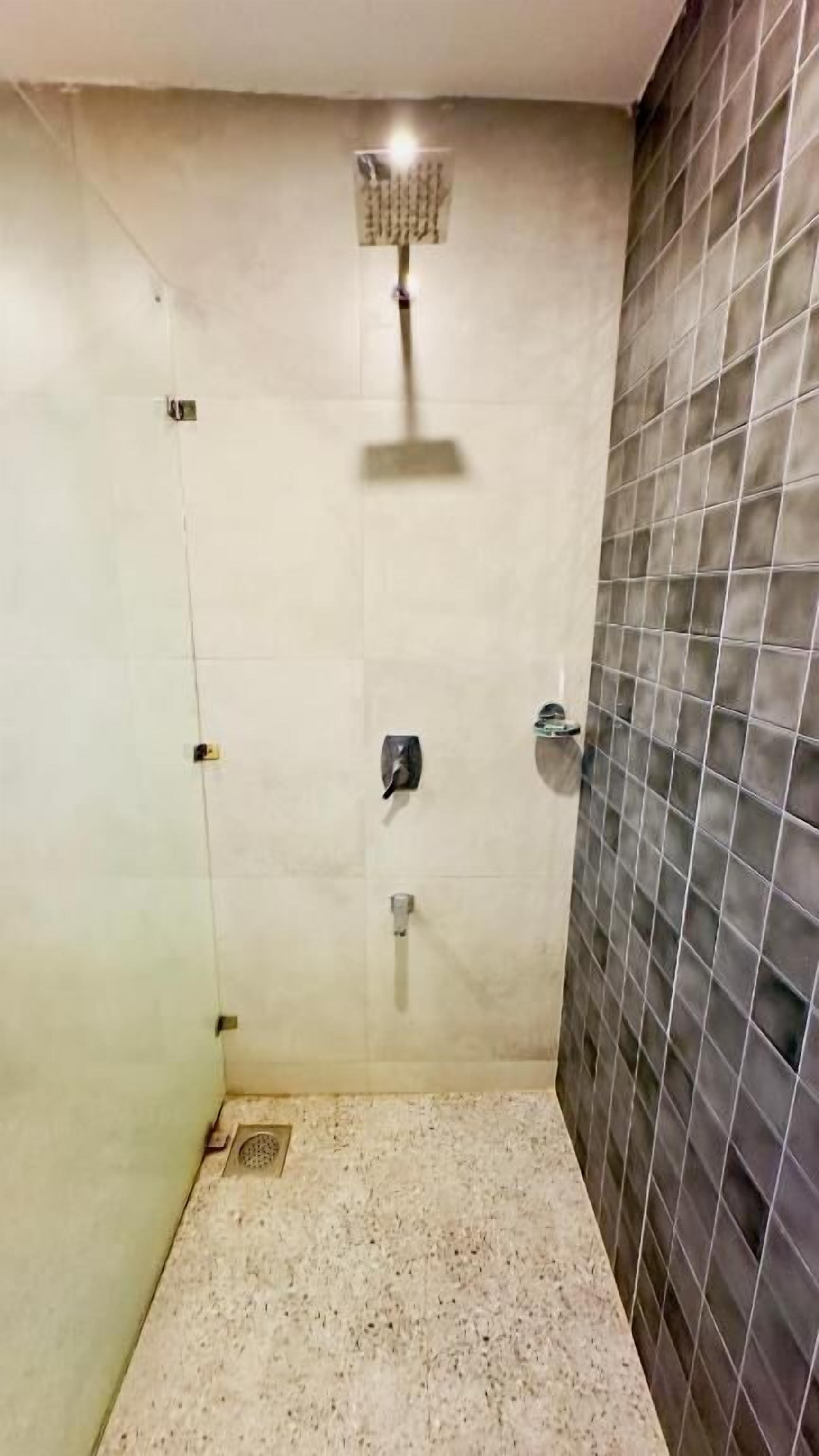 Un petit espace de douche carrelé, équipé d’un pommeau de douche à main et d’un robinet fixé au mur.