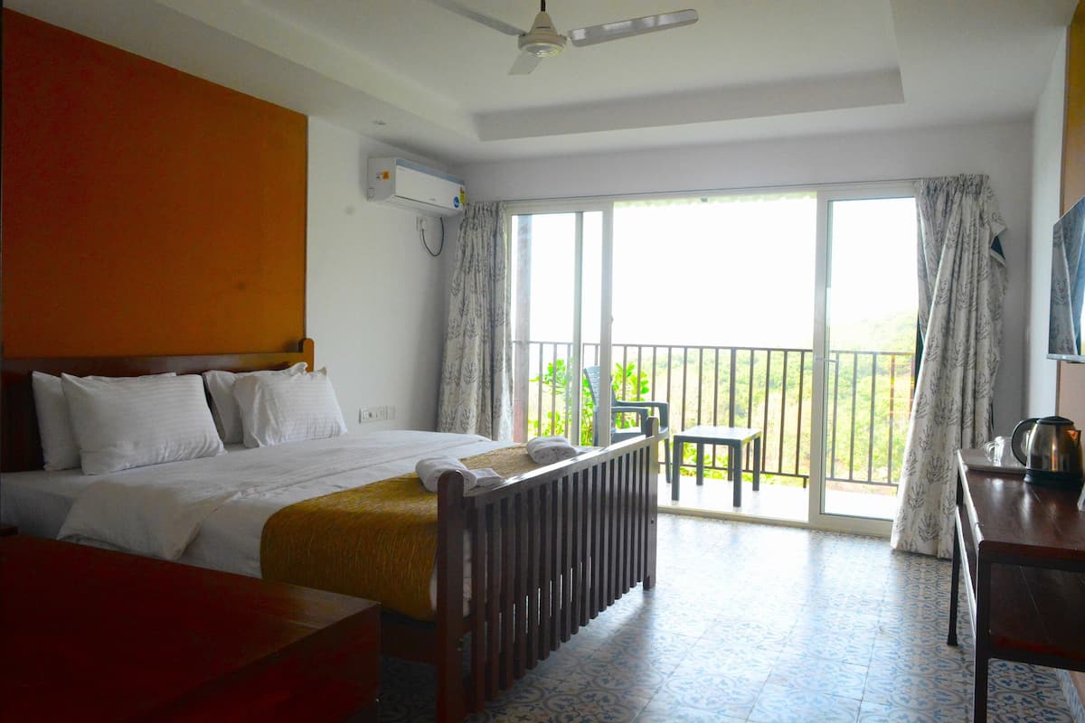 Habitación de hotel con cama, escritorio, silla, radiador y balcón con vistas a vegetación.