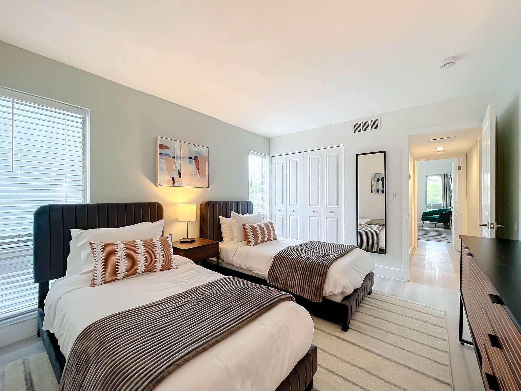 A modern bedroom with two beds, a large mirror, and a view into another room.