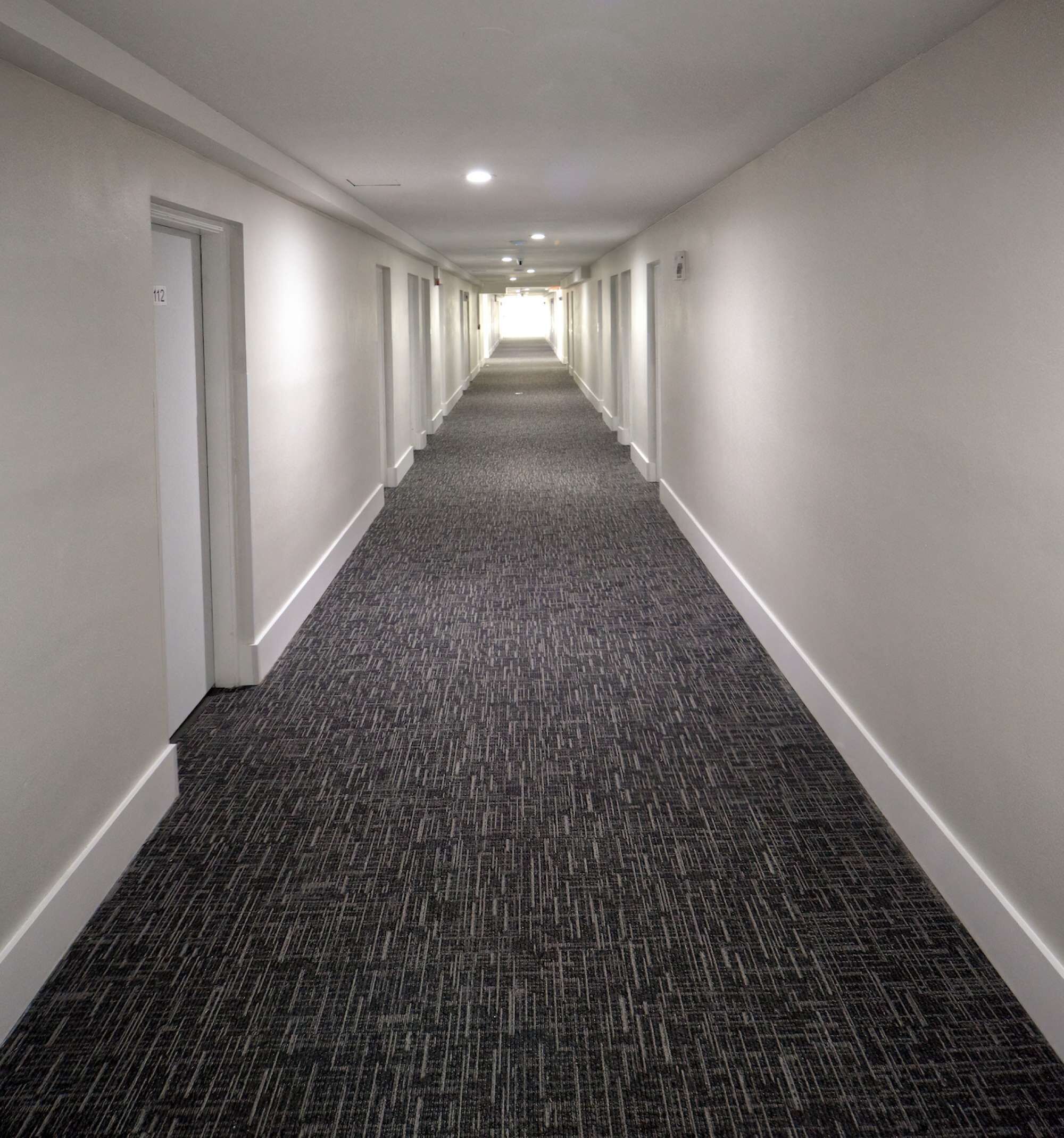 A long, narrow hallway with carpeted floors and white walls, leading to a bright end.