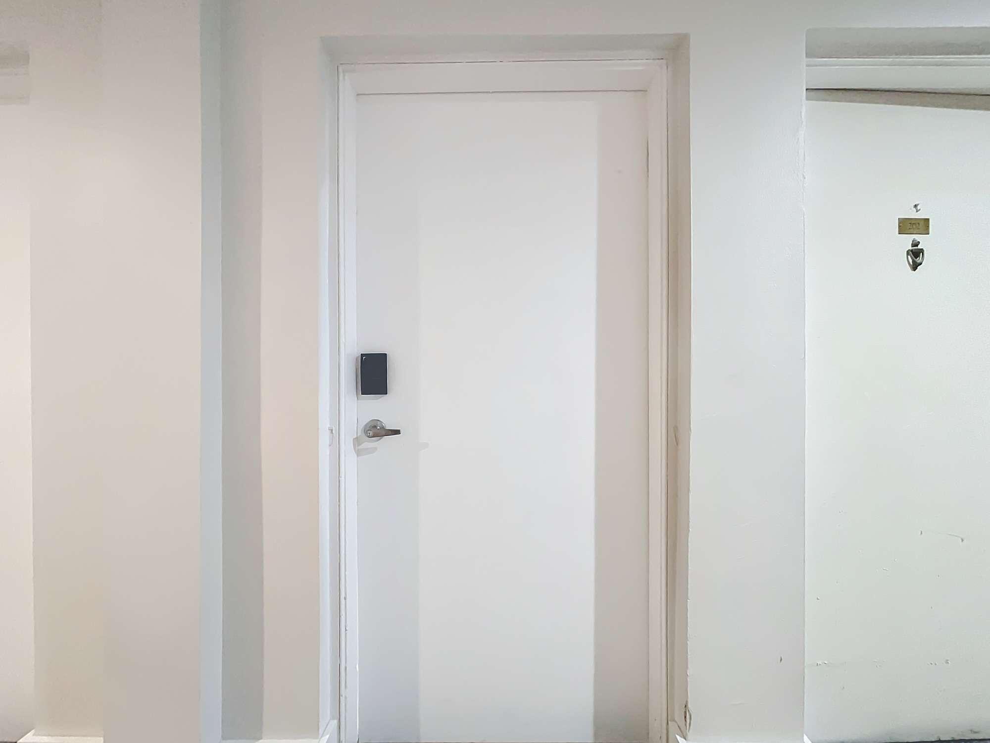 Uma porta branca com fechadura e olho de fechadura, um pequeno dispositivo eletrônico na porta e uma placa com o número da porta na parede ao lado.