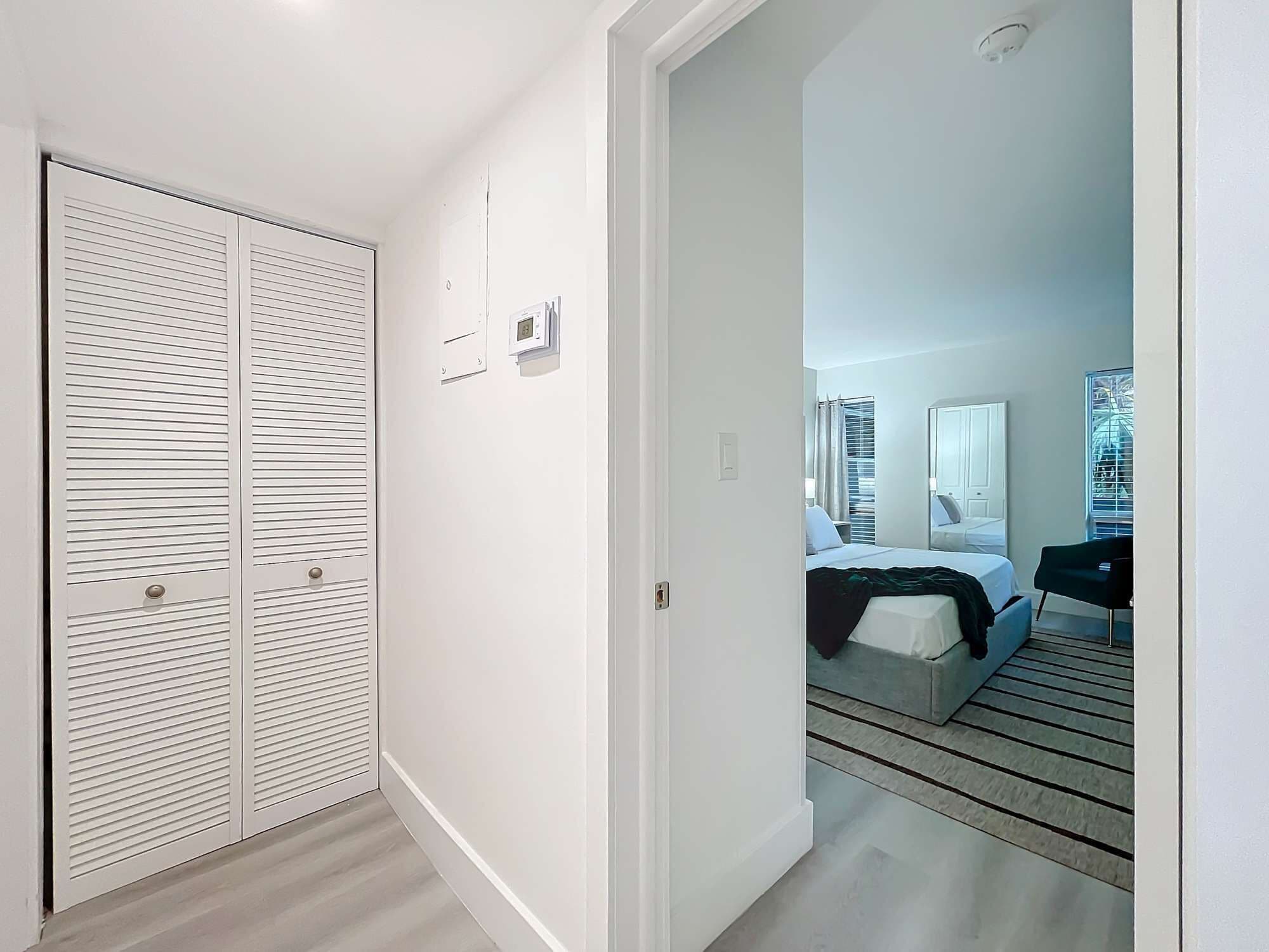 Habitación de hotel con cama, escritorio y una silla. Se ve un armario con puertas correderas y un termostato en la pared.