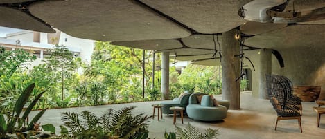 An indoor space with a modern design, featuring a seating area with a sofa and chairs, surrounded by lush greenery and a view of a building outside.