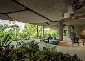 Un espace intérieur au design moderne, comprenant un coin salon avec un canapé et des fauteuils, entouré d’une végétation luxuriante et offrant une vue sur un bâtiment à l’extérieur.