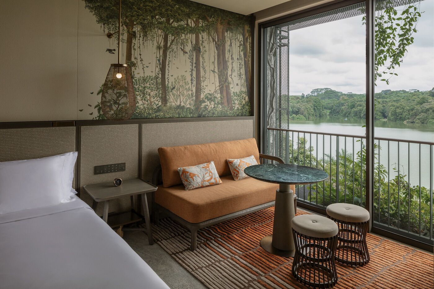 A modern hotel room with a large window overlooking a lake, a sofa with cushions, a small table, and a bed.