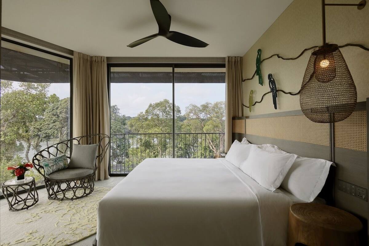 A hotel room with a large bed, a ceiling fan, a balcony with a chair, and a view of trees.