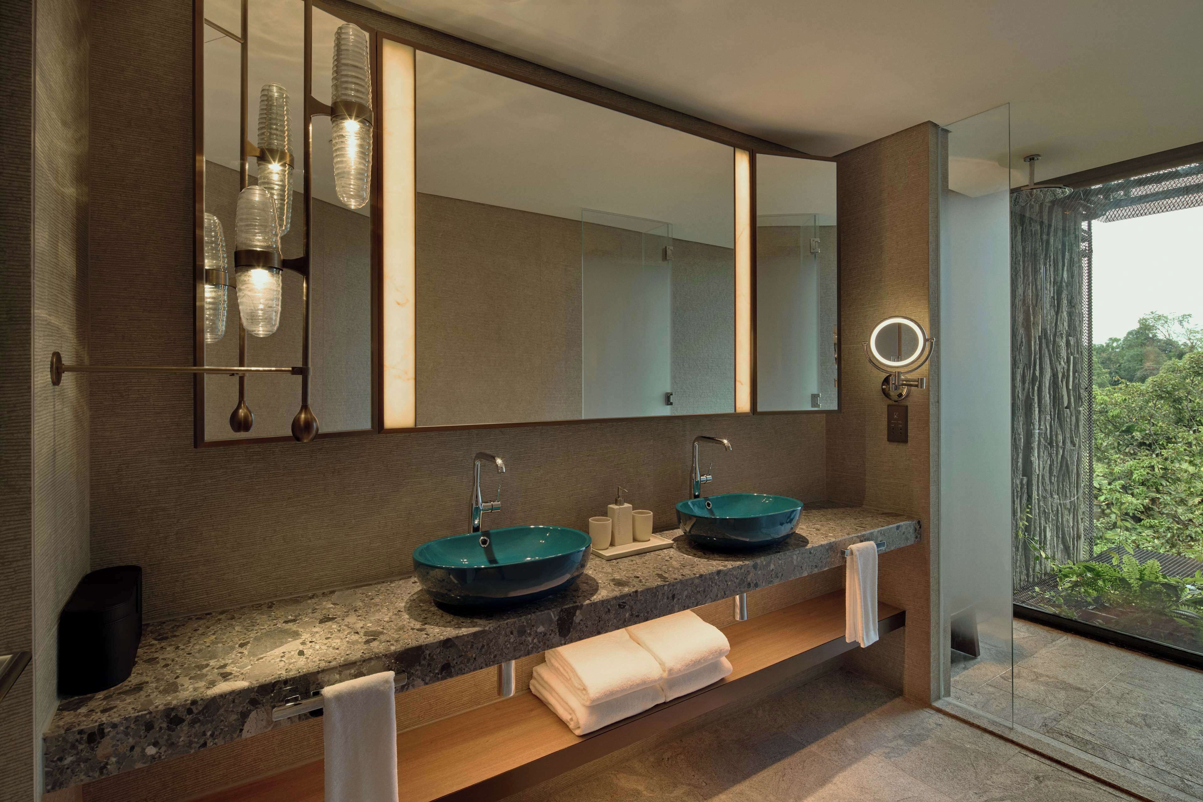 A modern bathroom with two sinks, a large mirror, and a view of greenery outside.