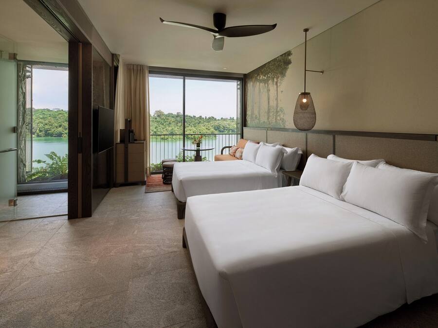 Habitación de hotel con dos camas, televisión, ventilador de techo y un amplio ventanal con vistas a un lago y vegetación.