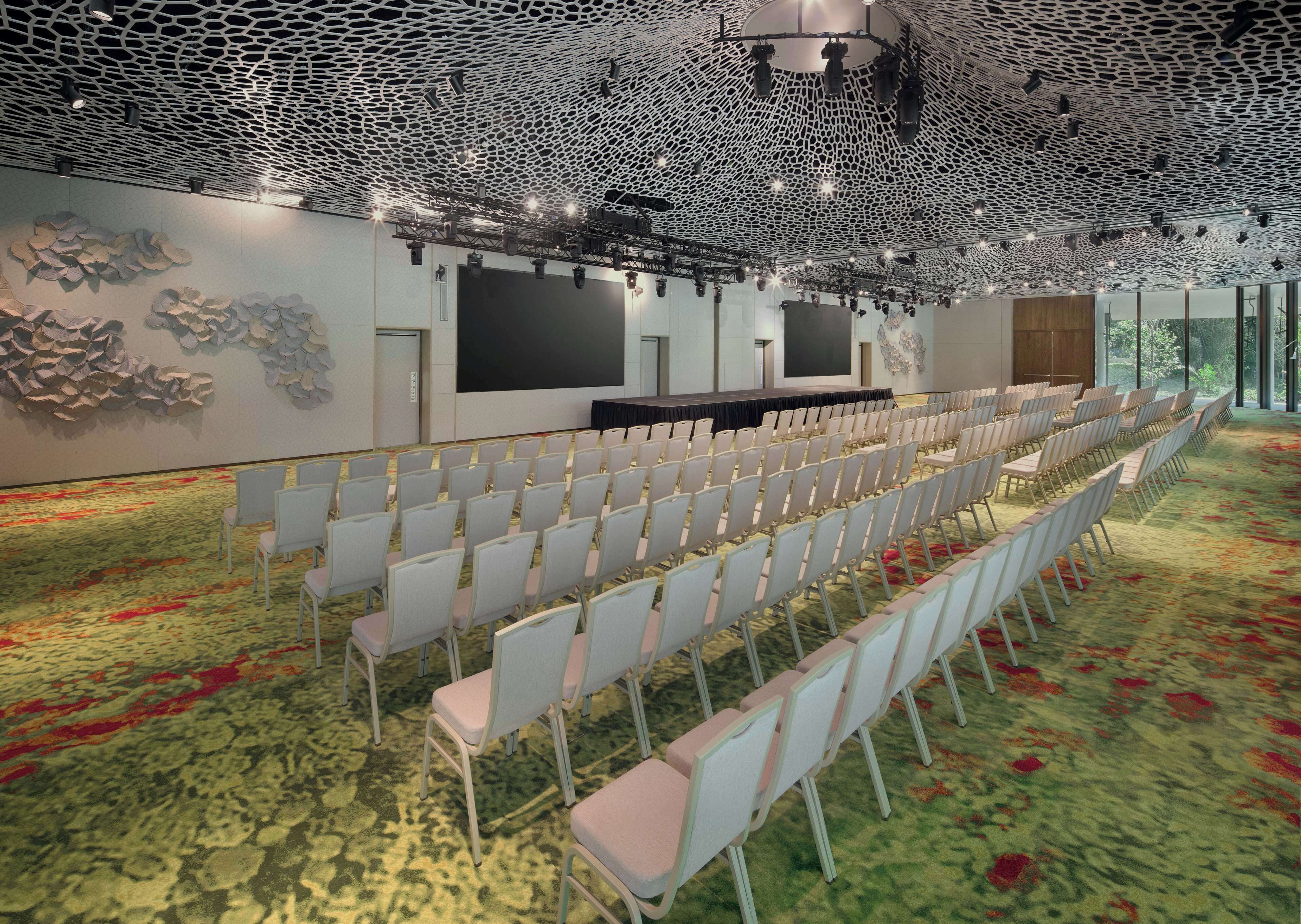 A conference room with rows of chairs, a stage with a black backdrop, and a ceiling with a unique lattice design.