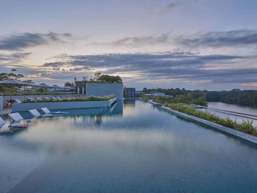 Una piscina infinita en la azotea con sillones de descanso, vista a un río y un edificio moderno con fachada de vidrio.
