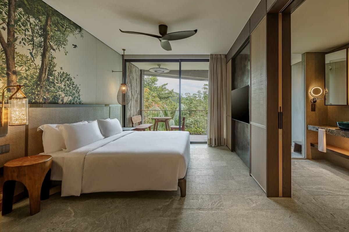 A modern hotel room with a large bed, a ceiling fan, a balcony with a view of greenery, and a bathroom with a mirror and sink.