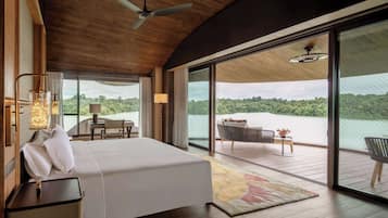 A bedroom with a bed, a desk, and a chair, a balcony with a view of a lake, and a ceiling fan.