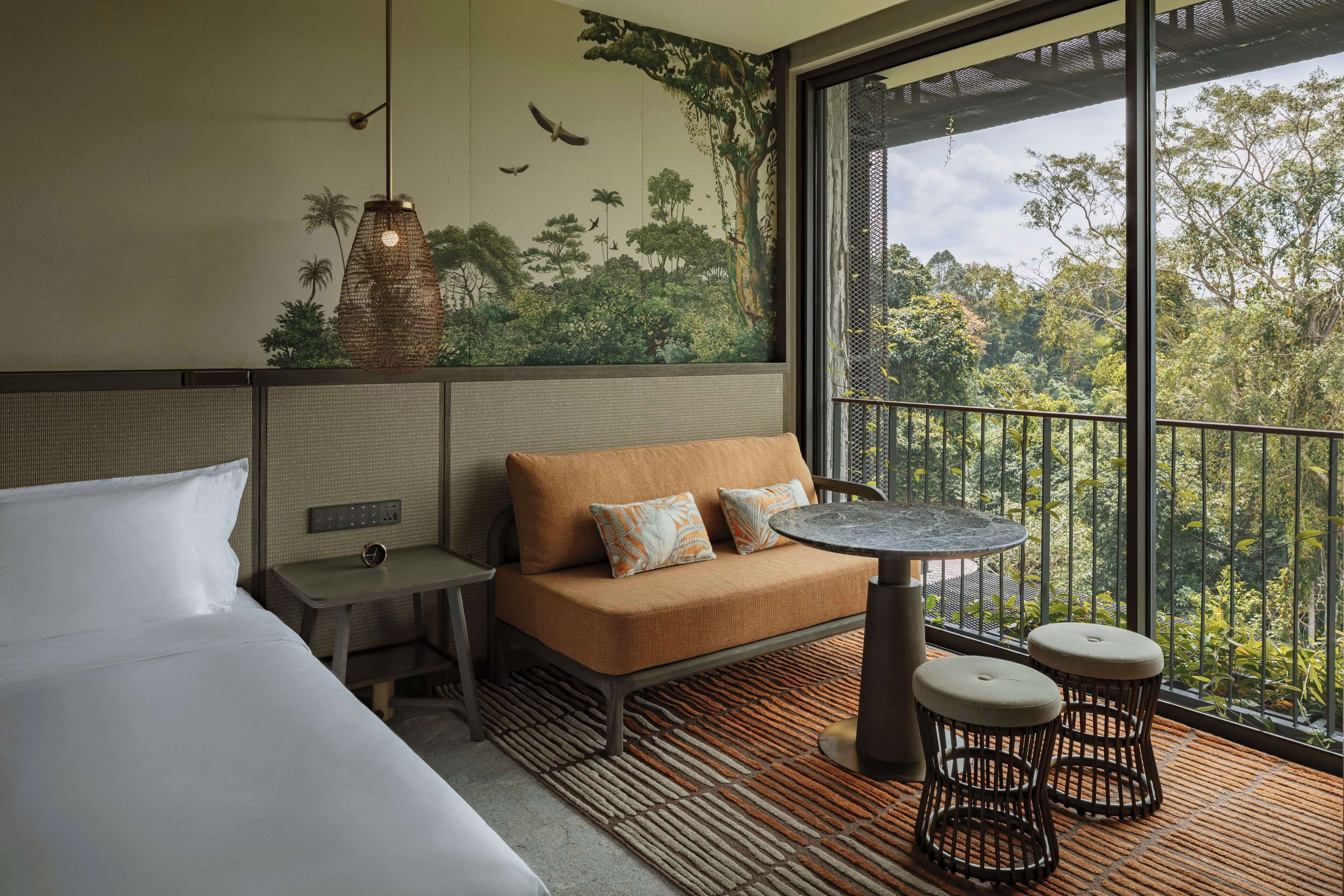 Habitación de hotel con cama, sofá, una mesita, dos taburetes, un mural en la pared y un balcón con vistas a árboles.