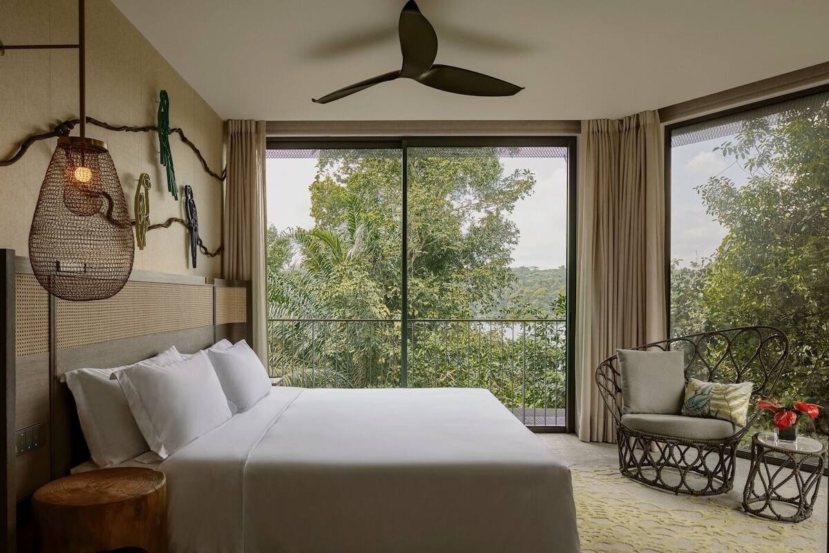 A bedroom with a large bed, a ceiling fan, a balcony with wicker furniture, and a view of greenery.