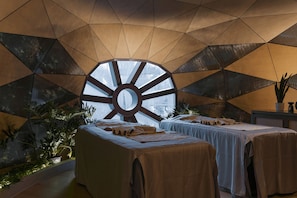 Una stanza con un design geometrico al soffitto, un'ampia finestra circolare, due letti con lenzuola bianche e una pianta sul lato.