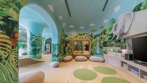 Zona de juegos infantiles con temática de selva, que incluye un gran mural de elefante, un espejo y un televisor.