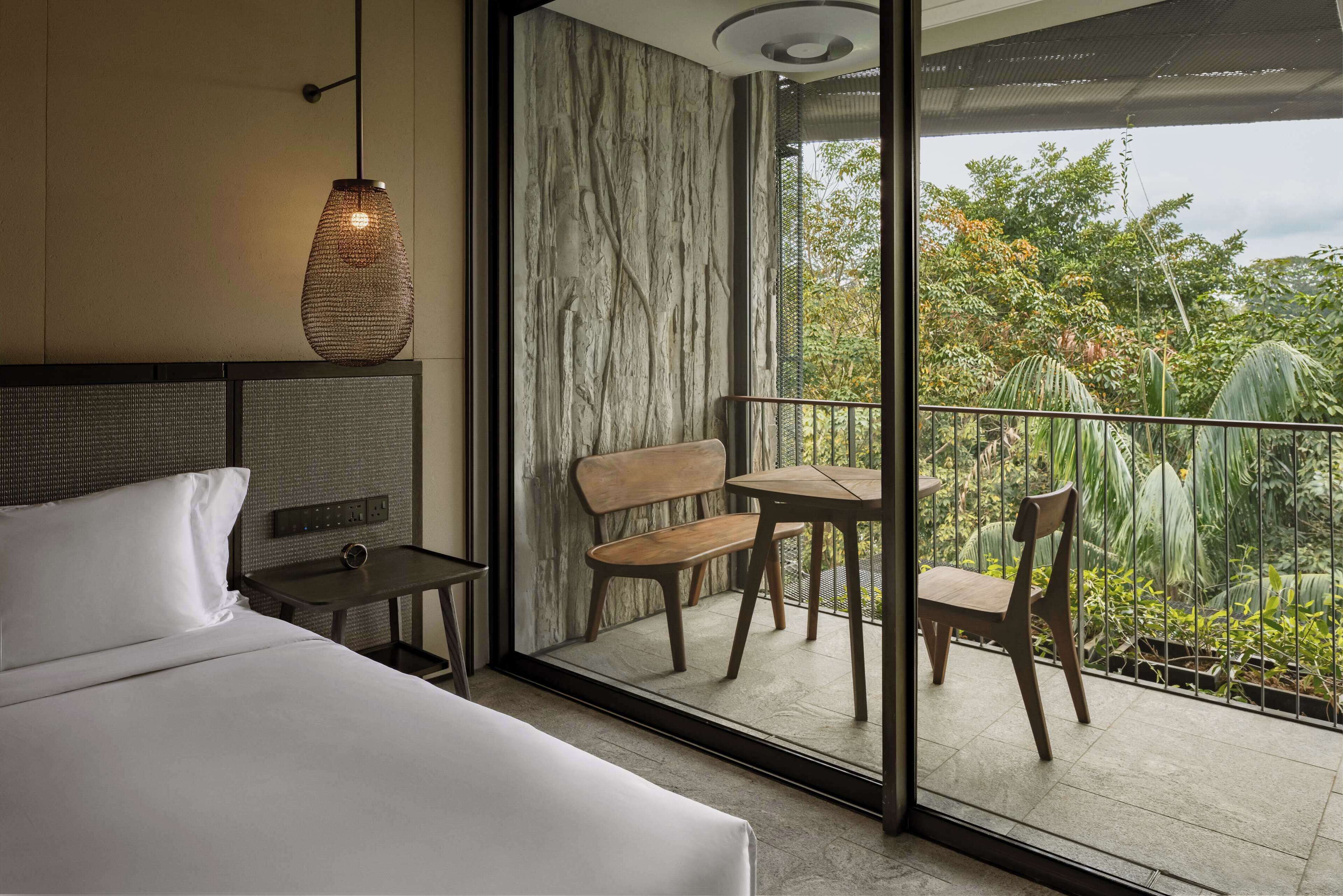 A hotel room with a bed, a small table with a chair, a hanging light fixture, and a balcony with a view of greenery.