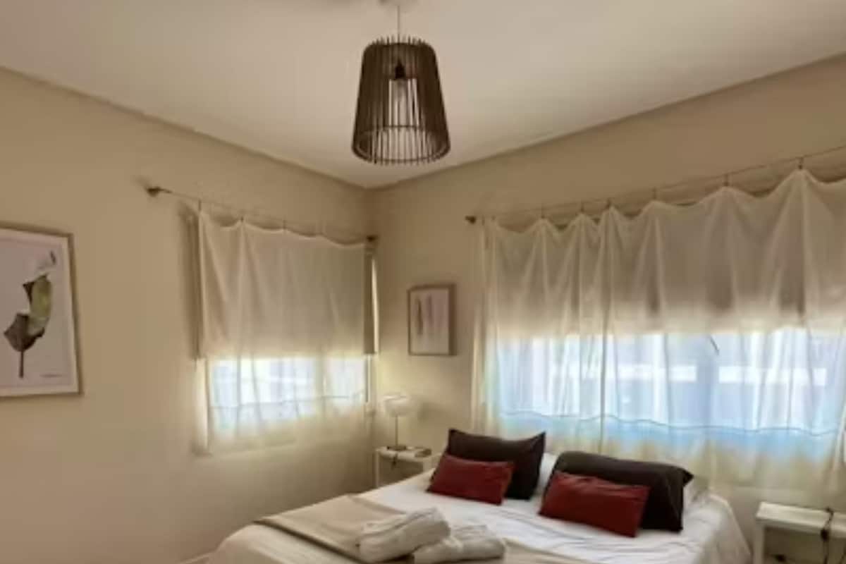 Ein Schlafzimmer mit einem Bett, weißen Vorhängen, einer Deckenleuchte und einem gerahmten Bild an der Wand.
