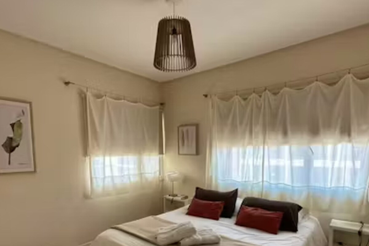 A bedroom with a bed, curtains, a bedside table, and a wall-mounted lamp.