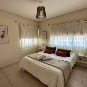 A bedroom with a bed, curtains, a bedside table, and a wall-mounted lamp. - Punto Playa (Villa Gesell)