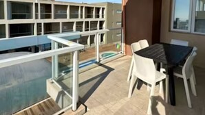 Une terrasse extérieure moderne, dotée d’une balustrade en verre, d’une piscine et d’un espace repas avec des chaises blanches et une table noire.