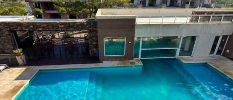 A rectangular swimming pool with clear blue water, surrounded by a stone deck. In the background, there is a multi-story building with balconies and a flat roof.
