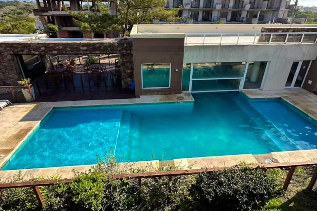 En rektangulär swimmingpool med klart, blått vatten, omgiven av en stenbelagd terrass. I bakgrunden syns en hög byggnad med flera våningar, balkonger och ett platt tak.