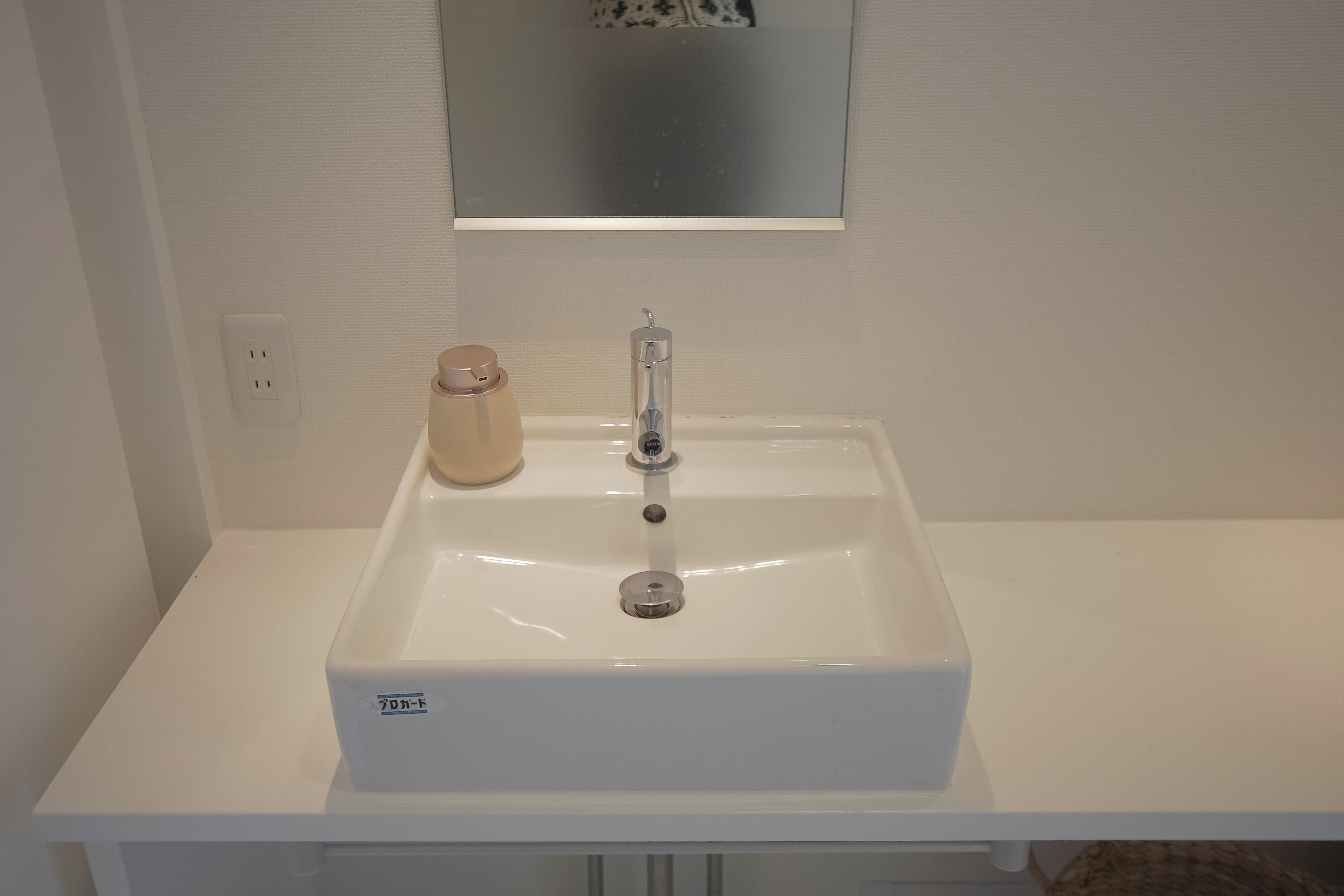 Un lavabo blanc dans une salle de bain, avec un miroir au-dessus, une petite bouteille et une prise électrique sur le mur.