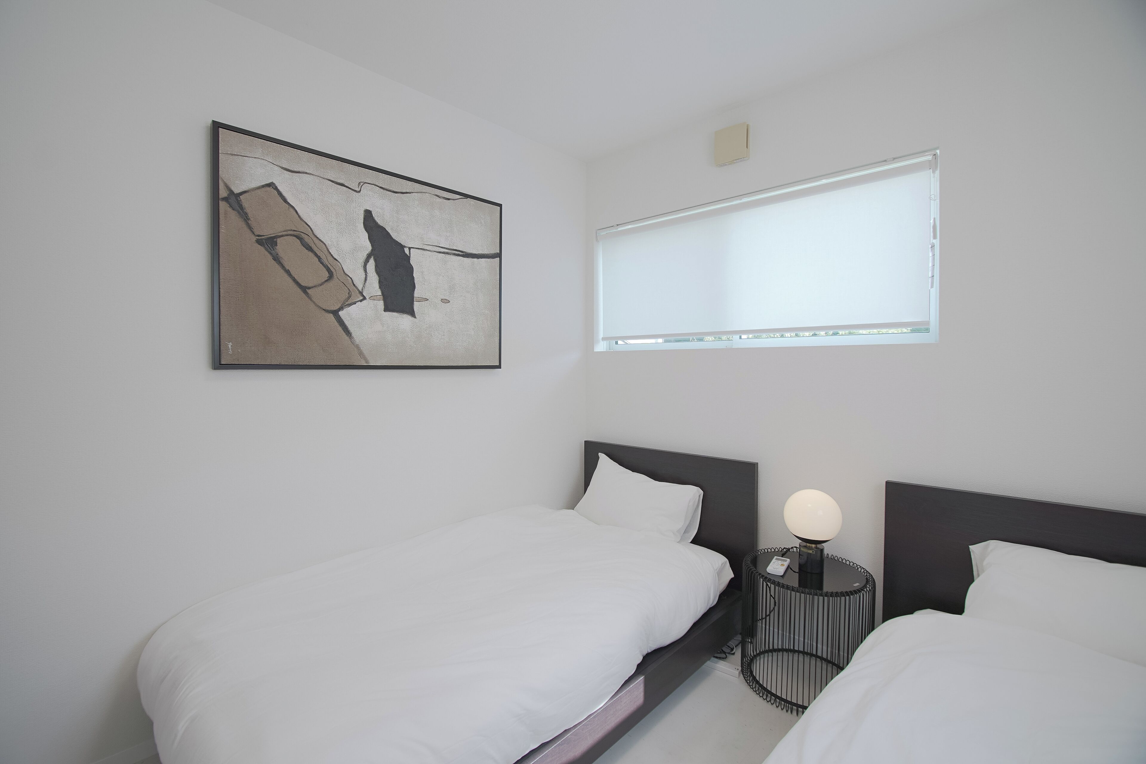 A hotel room with two beds, a bedside table with a lamp, a window with blinds, and a piece of abstract art on the wall.