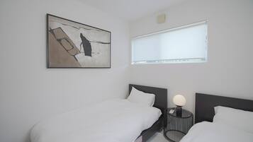 A hotel room with two beds, a bedside table with a lamp, a window with blinds, and a piece of abstract art on the wall.
