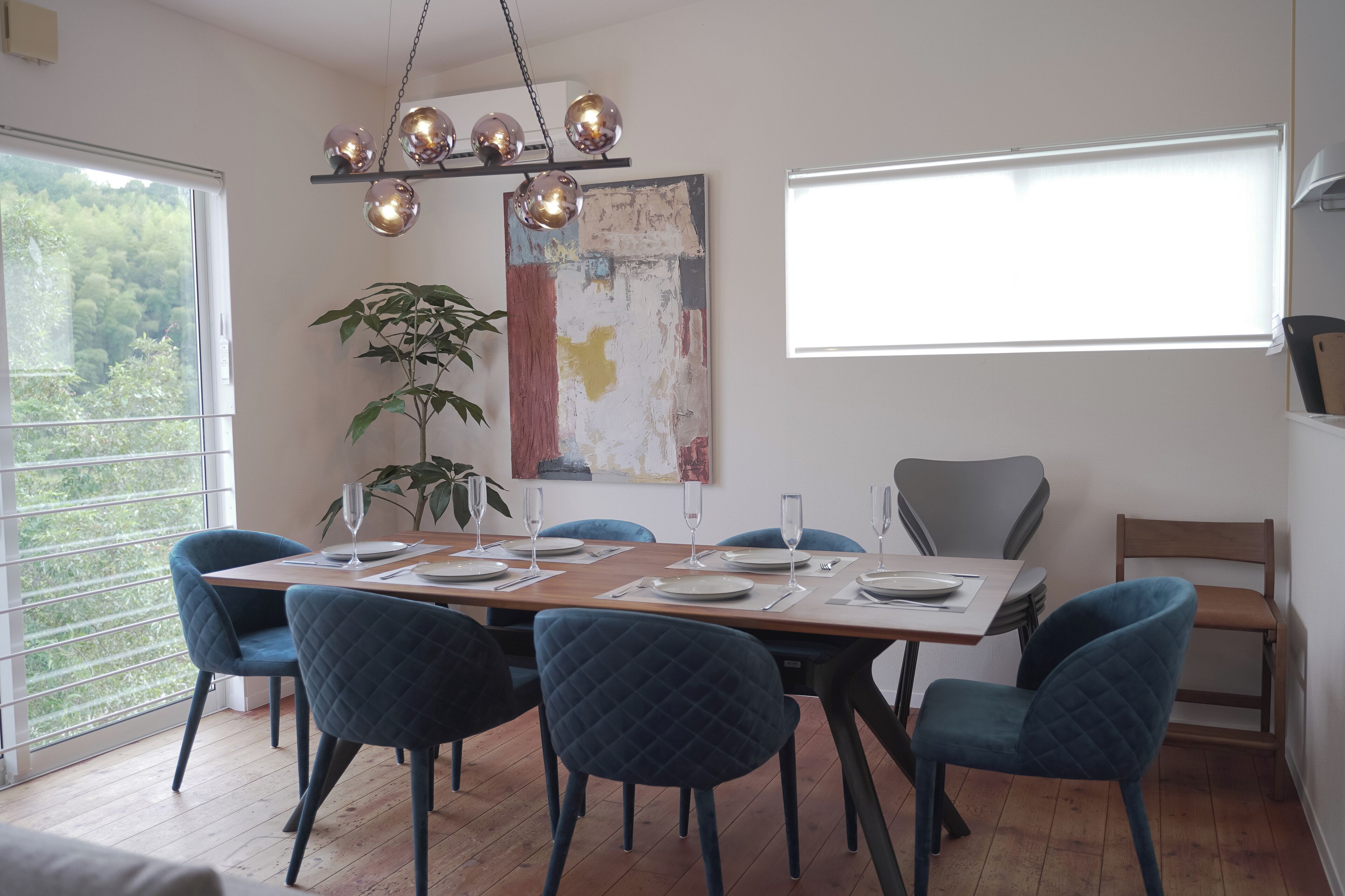 A dining area with a wooden table set for six, surrounded by six blue chairs. A large abstract painting and a potted plant are on the wall.