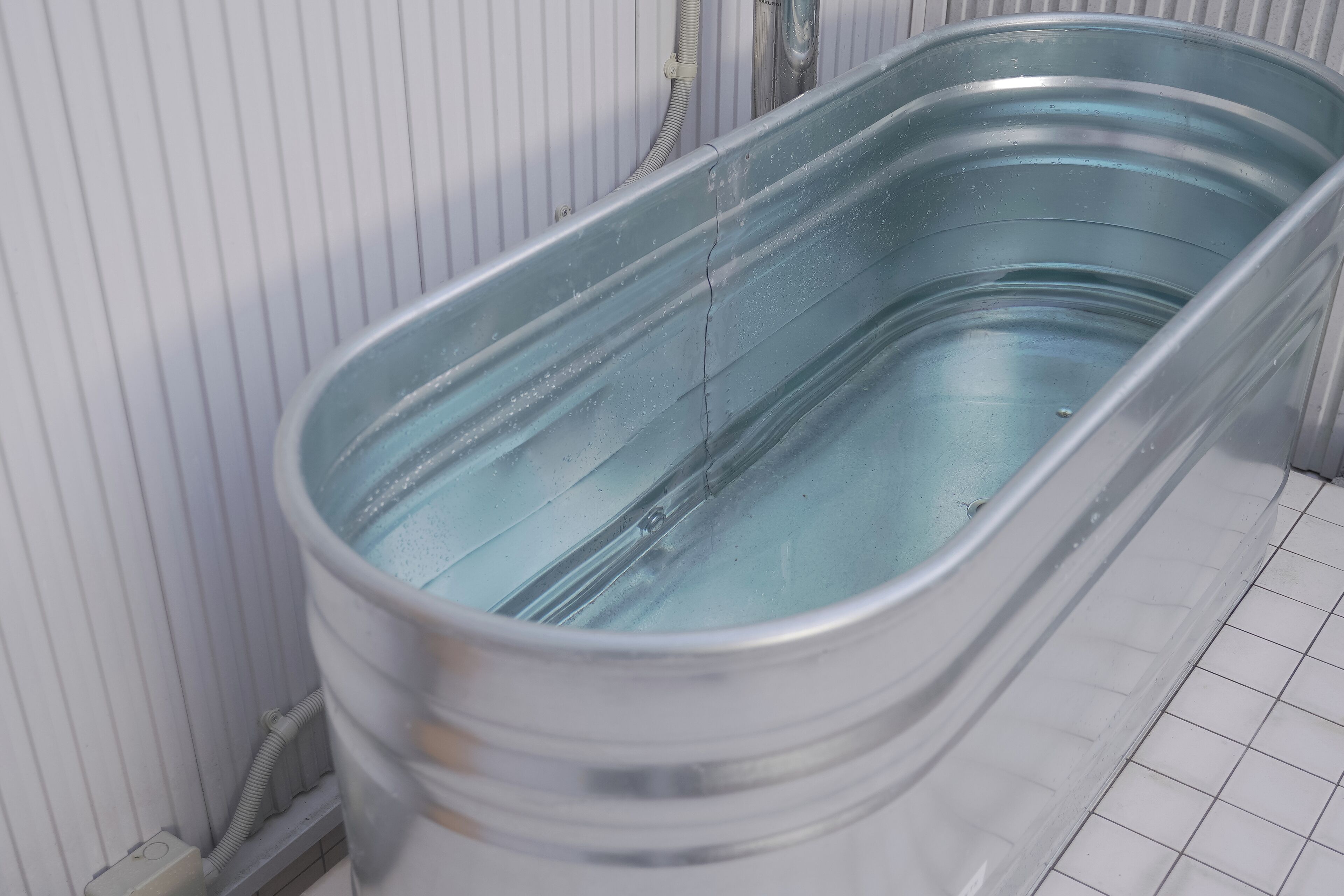 Una vasca da bagno in metallo piena d'acqua, un pavimento piastrellato di bianco e un muro bianco con una bocchetta di areazione.