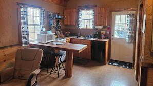 A cozy kitchen with wooden beams, a dining area with a chair and stools, and a view of the outdoors through a window.