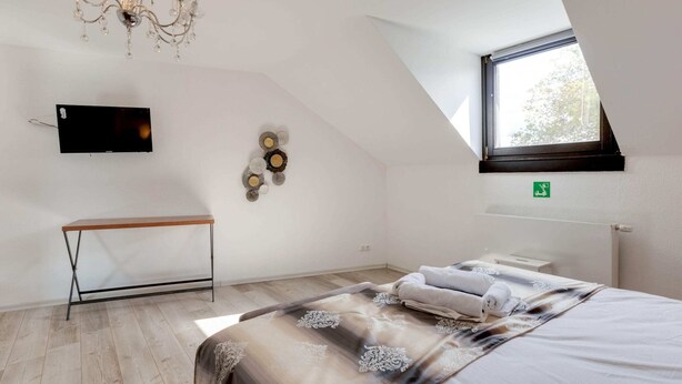 Une chambre avec un plafond en pente, un lustre, un téléviseur à écran plat, une table de chevet, un lit recouvert d’une couvre-lit à motifs et une fenêtre donnant sur les arbres.