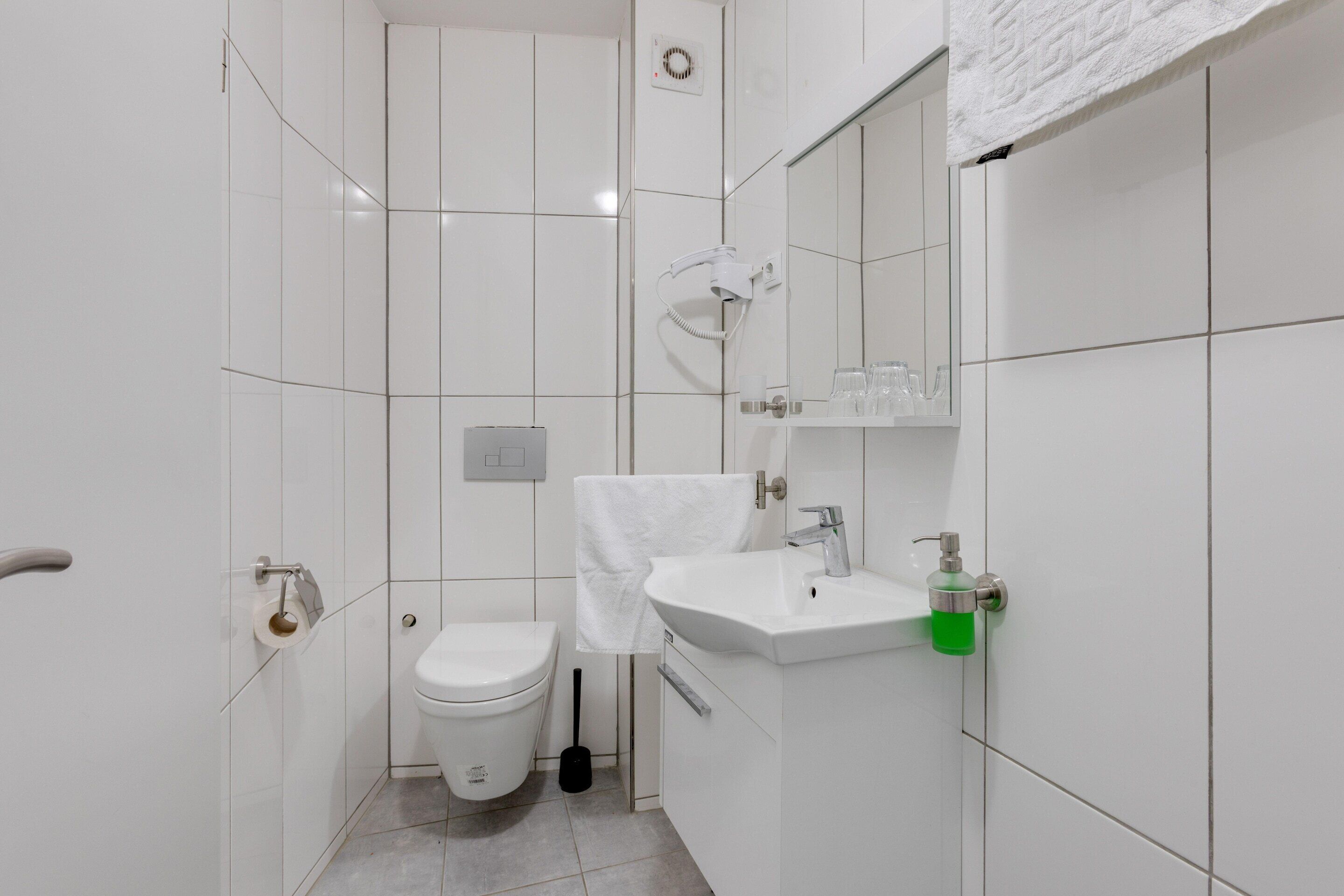 A small, modern bathroom with white tiles, a toilet, a sink, and a mirror.