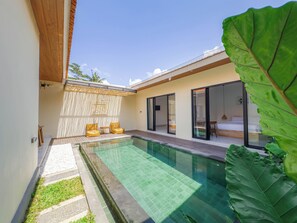 Un área de piscina moderna al aire libre, con una piscina cristalina, una zona de estar techada con cojines y una gran hoja verde en primer plano.