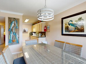 A dining area with a glass table and wicker chairs, a kitchen in the background, and a framed bird painting on the wall. - Beach Access Across the Street, Shared Heated Pool, Pet Friendly (Corpus Christi)