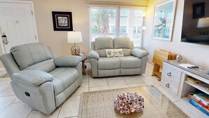 Ein Wohnzimmer mit einer Liegesofa, einem zusätzlichen Sofa, einem Beistelltisch mit Lampe, einer Holzkommode und einem an der Wand montierten Fernseher.