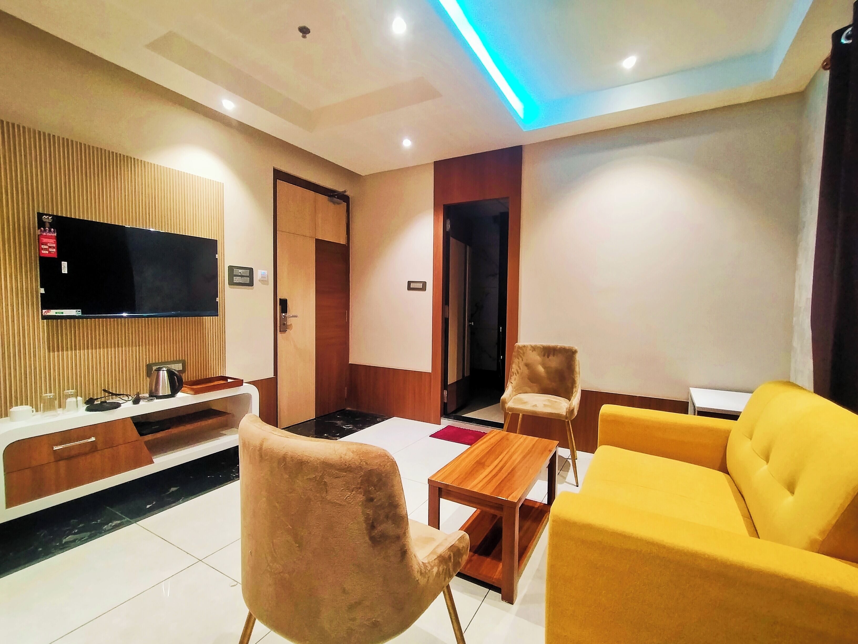 A modern hotel room with a flat-screen TV mounted on the wall, a wooden cabinet below, a yellow sofa, a small wooden table, and a pair of armchairs.
