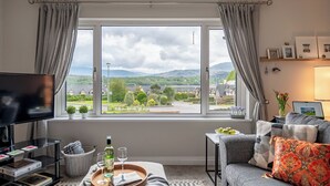 A living room with a TV, a sofa, a coffee table with books and glasses, a tray with a bottle and glasses, and a window with a view of a town and mountains. - 2 bedroom home in Kenmare with sea views to sleep 4 (Kenmare)