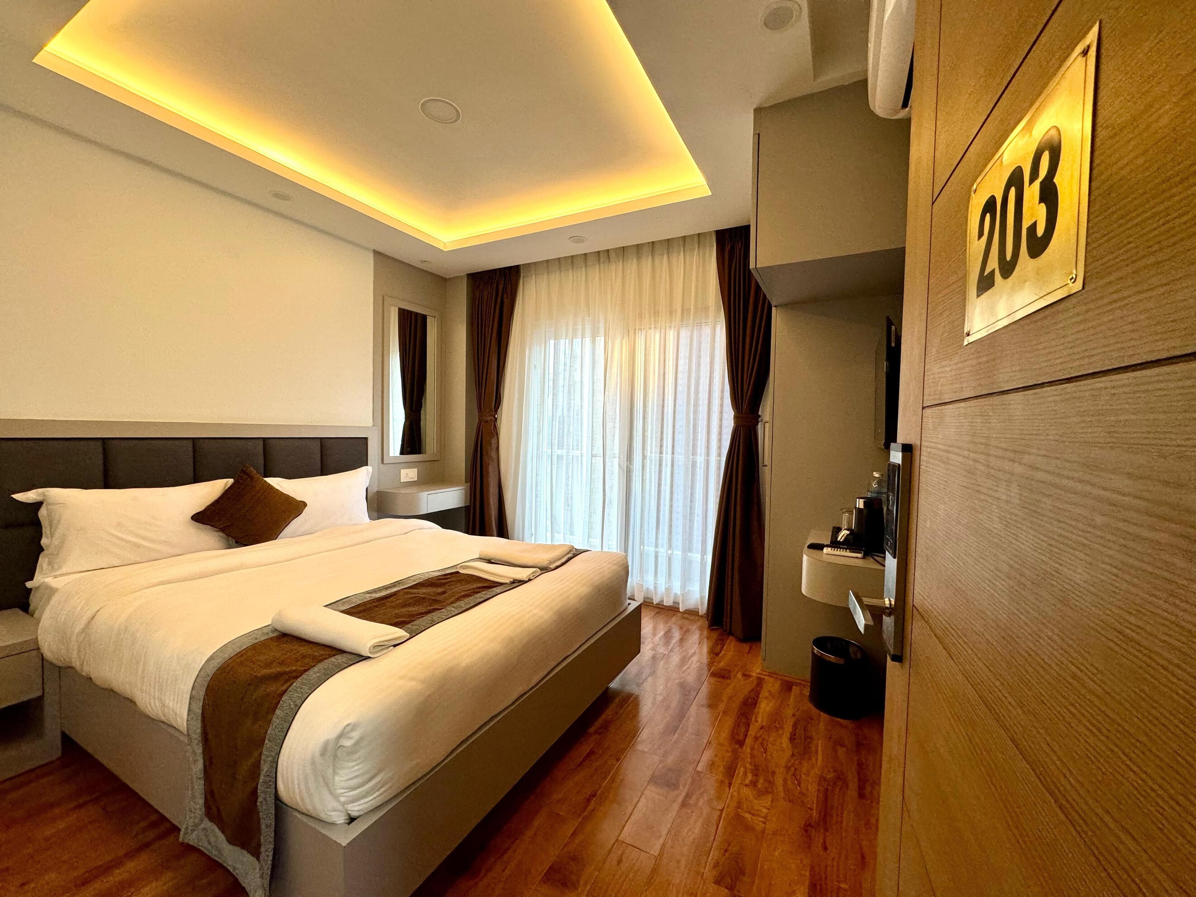 Une chambre d’hôtel moderne avec un grand lit, une table de chevet, un miroir, une porte en bois portant le numéro 203 et un plancher en bois.
