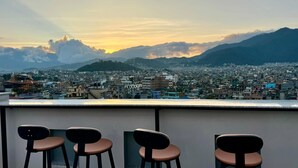 A rooftop bar with three stools overlooking a cityscape and mountains. - Hotel Alleyway Express (Kathmandu)