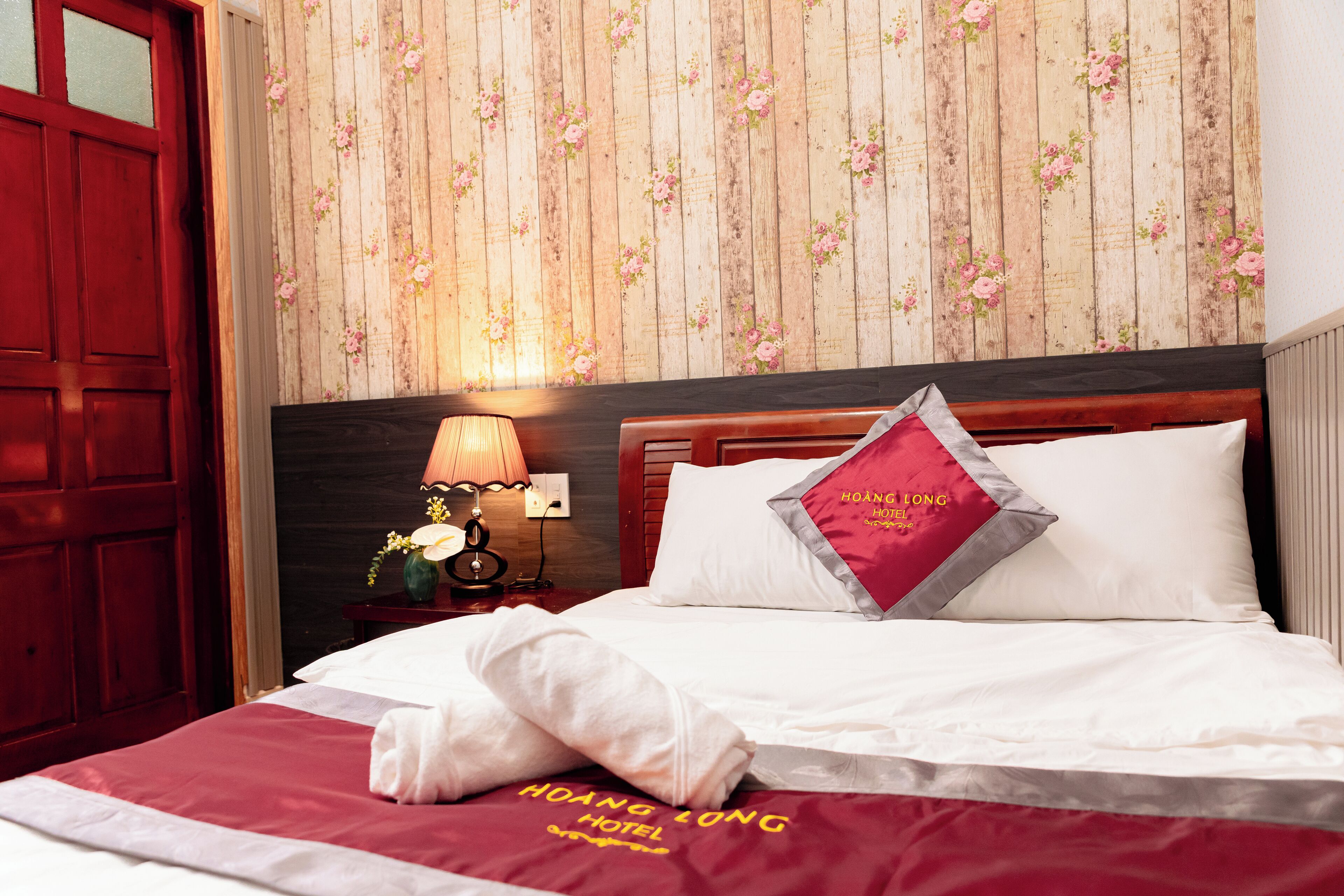 Quarto de hotel com cabeceira de madeira, cama com lençóis brancos, travesseiro vermelho com a marca do hotel e papel de parede floral.
